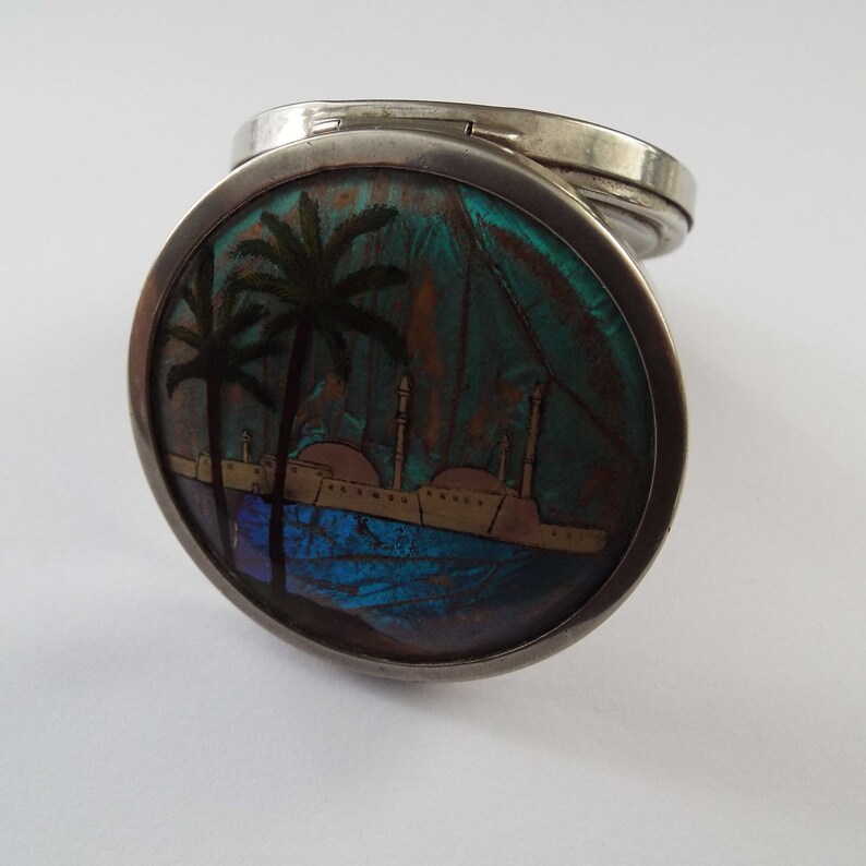 May include: A silver compact with a round, hinged lid. The lid is decorated with a colorful scene of palm trees and buildings reflected in a blue body of water. The scene is made from iridescent butterfly wings.