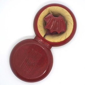 May include: A vintage, burgundy-colored compact with a matching lid. The open compact reveals a burgundy-colored, bat-wing shaped object nestled in a yellow, felt-like material. The compact is set against a white background.
