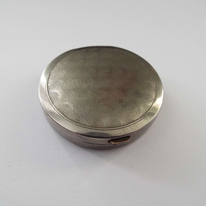 May include: A silver round compact with a wavy pattern on the lid.