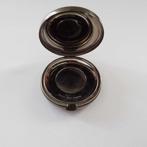 May include: A silver compact with a round mirror and a hinged lid. The compact has a black inscription that reads "WORLD WIDE PATENTS".
