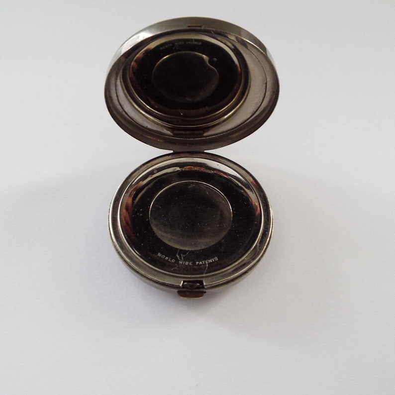 May include: A silver compact with a round mirror and a hinged lid. The compact has a black inscription that reads "WORLD WIDE PATENTS".