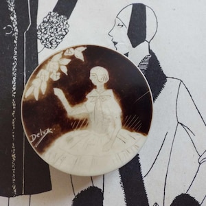Puede incluir: Broche vintage redondo, marrón y crema, con una ilustración detallada de una mujer con un vestido que alcanza una rama floral. El broche tiene la inscripción "Deluz". El fondo presenta una ilustración de moda en blanco y negro.