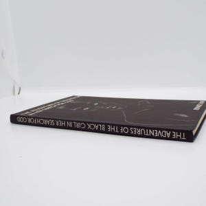 Puede incluir: Se muestra un libro negro titulado "THE ADVENTURES OF THE BLACK GIRL IN HER SEARCH FOR GOD". La portada del libro presenta una ilustraci&oacute;n en blanco y negro. El libro est&aacute; tumbado sobre una superficie blanca.
