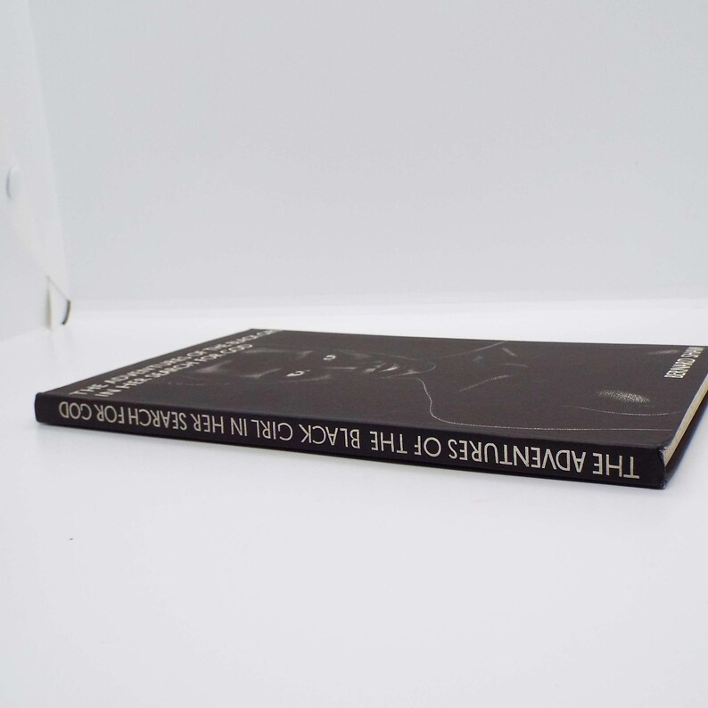 Puede incluir: Se muestra un libro negro titulado "THE ADVENTURES OF THE BLACK GIRL IN HER SEARCH FOR GOD". La portada del libro presenta una ilustraci&oacute;n en blanco y negro. El libro est&aacute; tumbado sobre una superficie blanca.