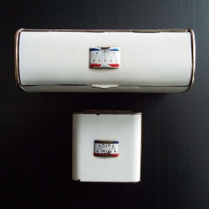 May include: Two white metal boxes, one rectangular and one square, with gold trim. Each box features a small, colorful emblem with a red, white, and blue design. The boxes are set against a dark background.