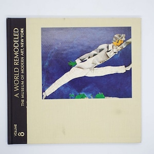 May include: A book titled "A World Remodeled" with a blue and white illustration of a person in a white suit with a green glove, jumping in the air. The book is volume 8 and is from The Museum of Modern Art, New York.