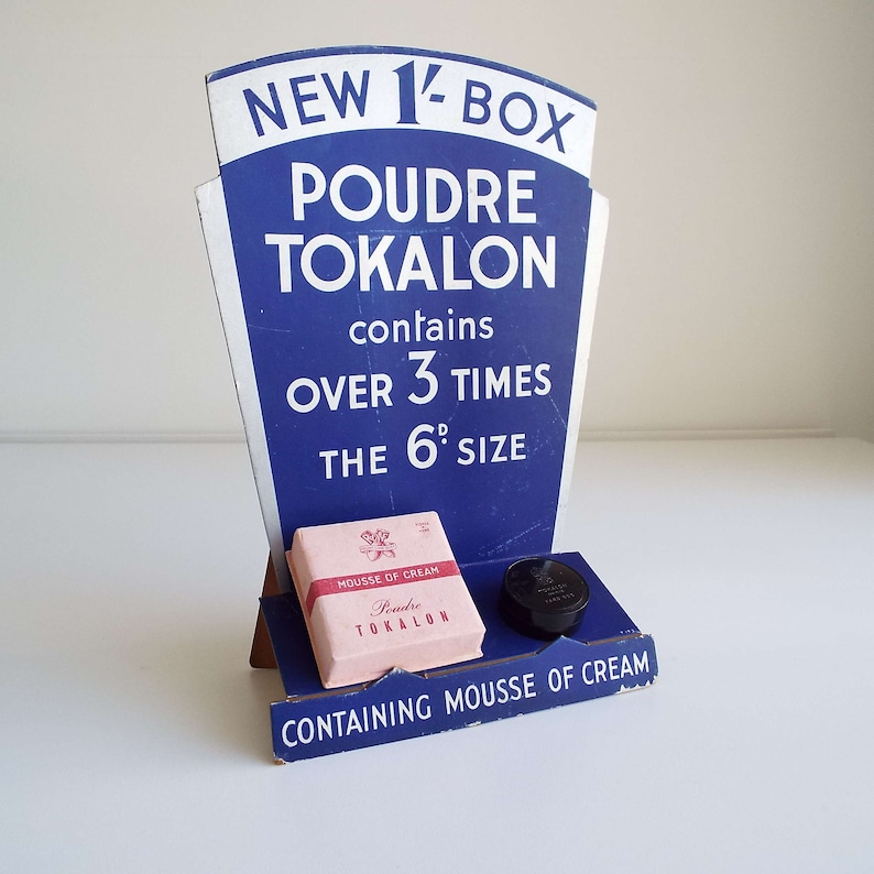 以下が含まれることがあります： 「Poudre Tokalon」という製品用の青と白の段ボール製ディスプレイスタンド。スタンドは、新しい1/2箱が6pサイズの3倍以上で、クリームのムースが含まれていることを宣伝しています。スタンドには、小さなピンクの箱に「Poudre Tokalon」という文字と、黒い丸い容器も付いています。
