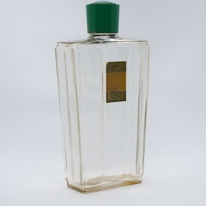 Op de afbeelding: Een heldere glazen fles met een groene plastic dop en een gouden etiket met groene rand. Het etiket heeft een decoratief ontwerp.