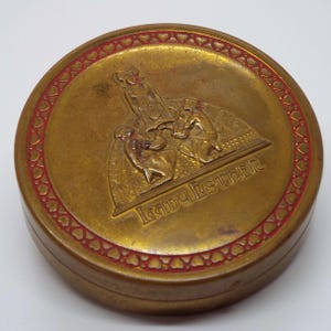 Puede incluir: Un compacto redondo de metal dorado con un borde de corazones rojos. El compacto presenta una imagen en relieve de una mujer con un vestido con dos figuras arrodilladas frente a ella. El texto "Lady Esther" está grabado en relieve en el compacto.