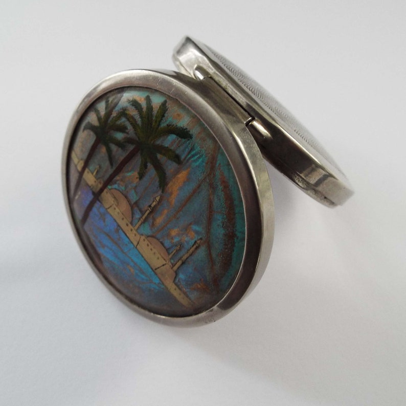 May include: A silver compact with a round, hinged lid. The lid features a colorful butterfly wing design with palm trees and a cityscape.