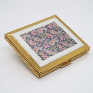 May include: A square, gold-toned compact with a decorative paisley design. The compact features a central panel with a navy blue background and pink and yellow paisley patterns. The frame is gold with a white border.