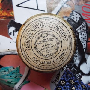May include: A vintage round powder compact with gold edging. The label features French text, including "Poudre de Riz". The compact is beige with black lettering and a decorative border. The text includes "Fabrique Speciale de Produits" and "Pour la Beaute des Dames".