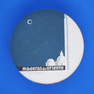 May include: A round, vintage coaster with a blue and white design. The coaster features a stylized illustration of a building with the text "maderas de oriente" and "MYRURGIA". The background is a gradient of blue.