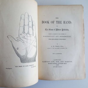 May include: Open book with the title "The Book of the Hand" and an illustration of a hand with lines and markings. The book's subtitle is "The Science of Modern Palmistry". Published in London in 1867.