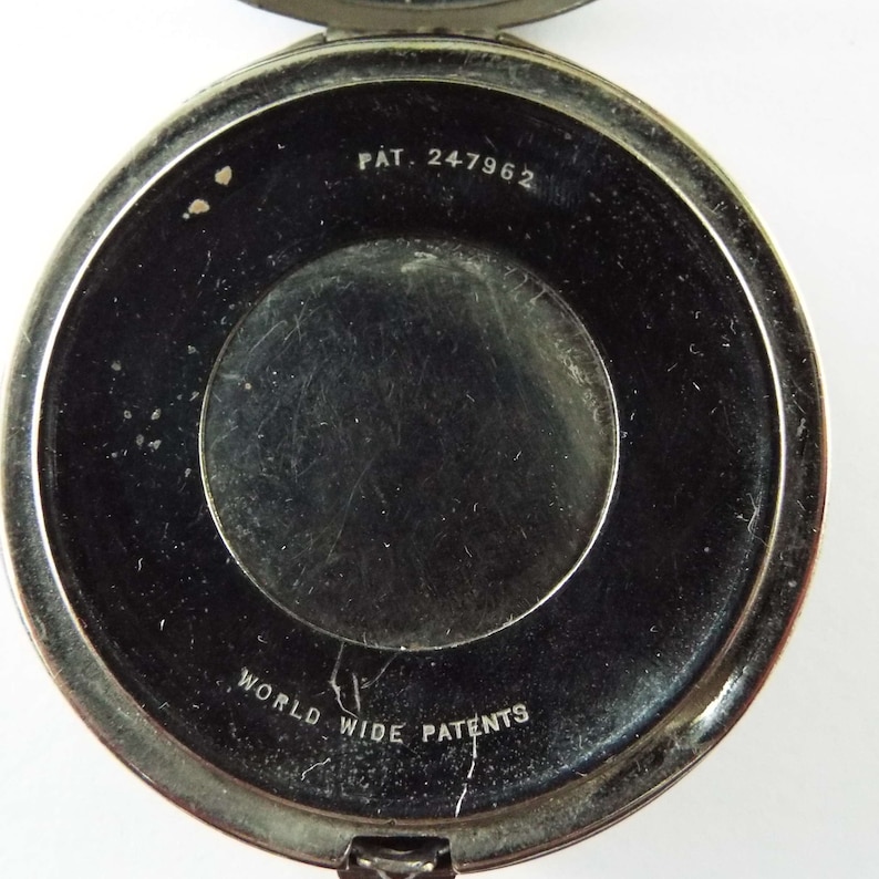 May include: The underside of a silver compact with the text "PAT. 247962" and "WORLD WIDE PATENTS" inscribed.