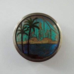 May include: A round silver brooch with a colorful scene of a city with palm trees and a blue body of water. The scene is made from iridescent butterfly wings.