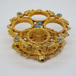 Peut inclure: Support décoratif en métal doré avec trois ouvertures circulaires et un pilier central. Le support est orné de fausses perles et de pierres de couleur turquoise.