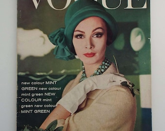 May include: Vintage Vogue magazine cover featuring a woman wearing a green hat and a cream-colored coat. The cover includes text that reads "VOGUE" and "FEBRUARY, 1961". The woman is wearing a turquoise necklace and white gloves.