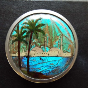 May include: A silver circular brooch with a blue and green iridescent background. The brooch features a painted scene of a city with palm trees and white buildings.
