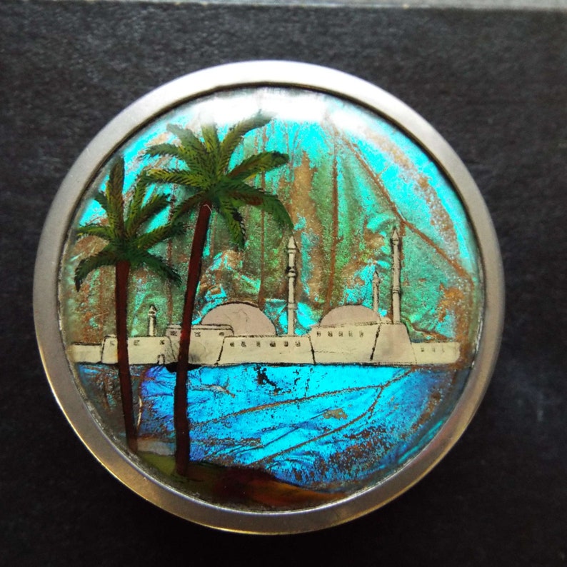 May include: A silver circular brooch with a blue and green iridescent background. The brooch features a painted scene of a city with palm trees and white buildings.