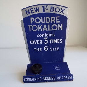 以下が含まれることがあります： 青と銀の段ボール製のディスプレイスタンドに「NEW 1/2 BOX POUDRE TOKALON contains OVER 3 TIMES THE 6d SIZE」と宣伝されています。スタンドには、製品の箱を置くための小さな切り抜きがあります。「CONTAINING MOUSSE OF CREAM」というテキストがスタンドの下部に印刷されています。