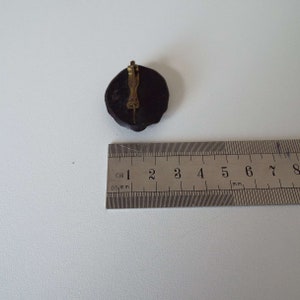 May include: A round brooch in black and gold with a decorative design. The brooch has a metal clasp on the back.