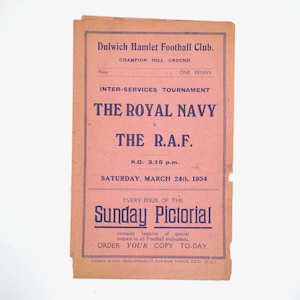 Könnte beinhalten: Vintage Fußballspielprogramm für den Dulwich Hamlet Football Club. Das rosa Papier zeigt dunkelblauen Text, darunter "The Royal Navy v. The R.A.F." und das Datum "Samstag, 24. März 1934." Es wirbt auch für den Sunday Pictorial.