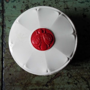 Op de afbeelding: Een witte, ronde container met een rood, cirkelvormig embleem in het midden. Het embleem heeft een gedetailleerd insectenontwerp. De container heeft een bloembladachtig ontwerp rond de rand.