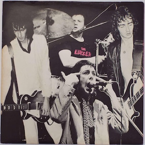 Könnte beinhalten: Schwarz-Weiß-Foto einer Rockband, die auf der Bühne auftritt. Der Leadsänger in einer gestreiften Jacke zeigt mit dem Finger auf seinen Kopf, während er in ein Mikrofon singt. Andere Bandmitglieder spielen Gitarre und Schlagzeug. Ein Mitglied trägt ein T-Shirt mit "The Lurker".