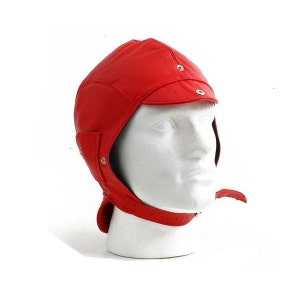 Puede incluir: Un casco de aviador de cuero rojo brillante con orejeras y una correa para la barbilla. El casco tiene una visera y se asegura con remaches metálicos. La correa de la barbilla es ajustable. El casco se muestra en una cabeza de maniquí blanca.