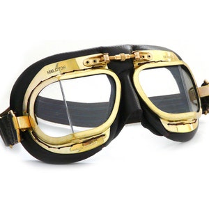 Könnte beinhalten: Schwarze Leder-Fliegerbrille mit goldfarbenen Metallrahmen und klaren Gläsern. Die Brille hat ein Band und der Text "HALCYON" ist auf dem Rahmen aufgedruckt.