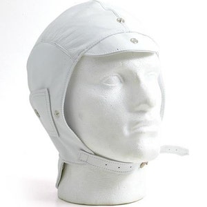 以下が含まれることがあります： ストラップとバックル留め具が付いた白い革製のパイロットハット。帽子にはバイザーと銀色のスナップが2つ付いています。