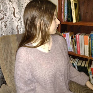 Pu&ograve; includere: Una donna indossa un maglione di colore lilla chiaro e soffice con una collana d'argento. &Egrave; seduta su una sedia di fronte a una libreria piena di libri.