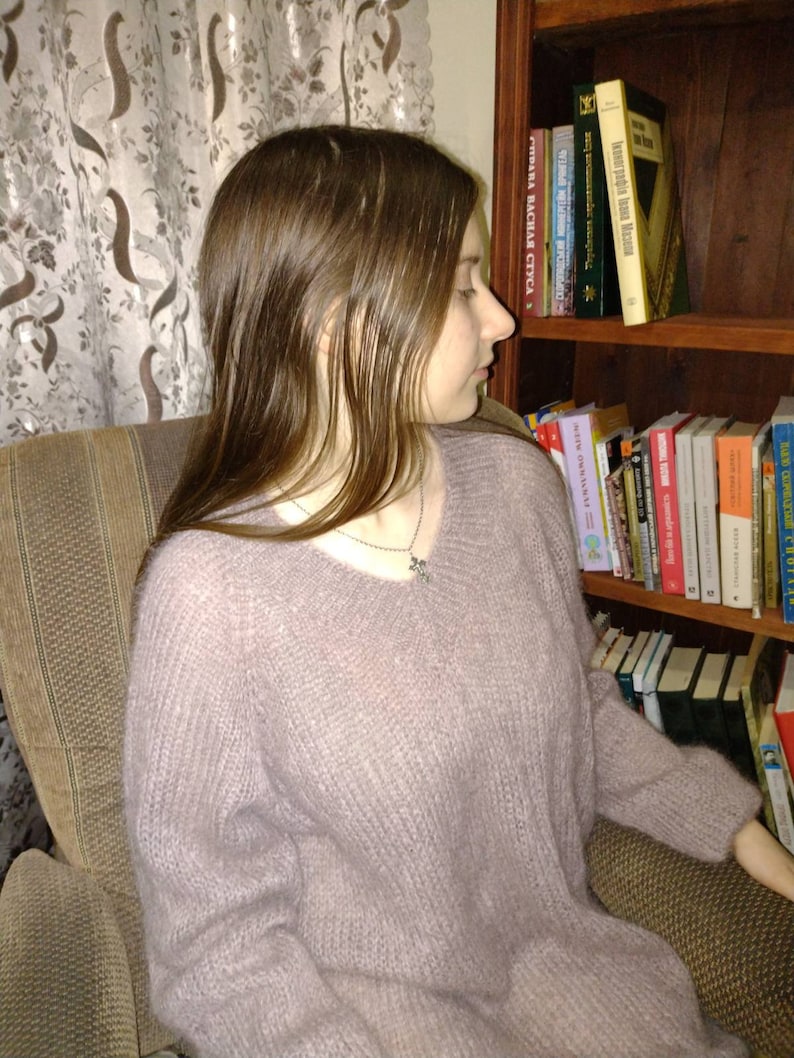 Pu&ograve; includere: Una donna indossa un maglione di colore lilla chiaro e soffice con una collana d'argento. &Egrave; seduta su una sedia di fronte a una libreria piena di libri.