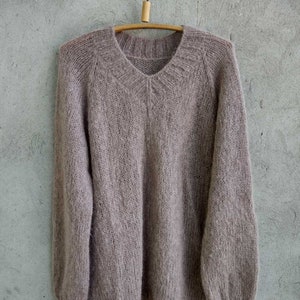 Pu&ograve; includere: Un maglione di lana morbido, di colore marrone chiaro, con scollo a V e maniche lunghe. Il maglione &egrave; appeso a un appendiabiti di legno su uno sfondo grigio.