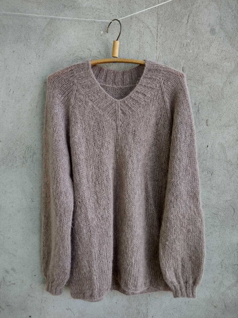 Pu&ograve; includere: Un maglione di lana morbido, di colore marrone chiaro, con scollo a V e maniche lunghe. Il maglione &egrave; appeso a un appendiabiti di legno su uno sfondo grigio.