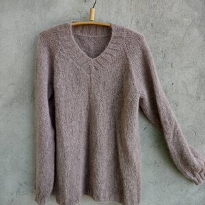 Pu&ograve; includere: Un maglione di maglia morbido color malva con scollo a V e maniche lunghe appeso a un appendiabiti di legno.