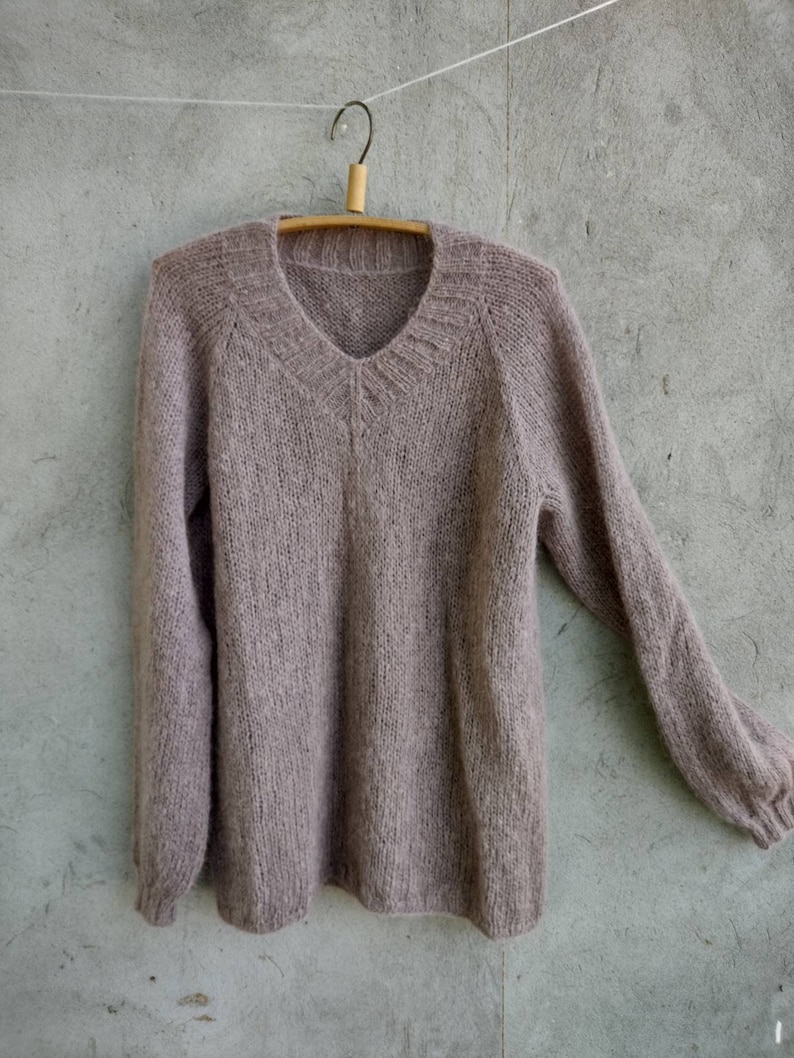 Pu&ograve; includere: Un maglione di maglia morbido color malva con scollo a V e maniche lunghe appeso a un appendiabiti di legno.