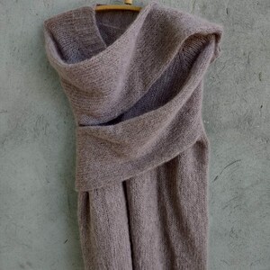 Pu&ograve; includere: Un maglione di maglia morbida color malva con collo a lupetto e un design drappeggiato. Il maglione &egrave; appeso a un appendiabiti di legno su uno sfondo grigio.