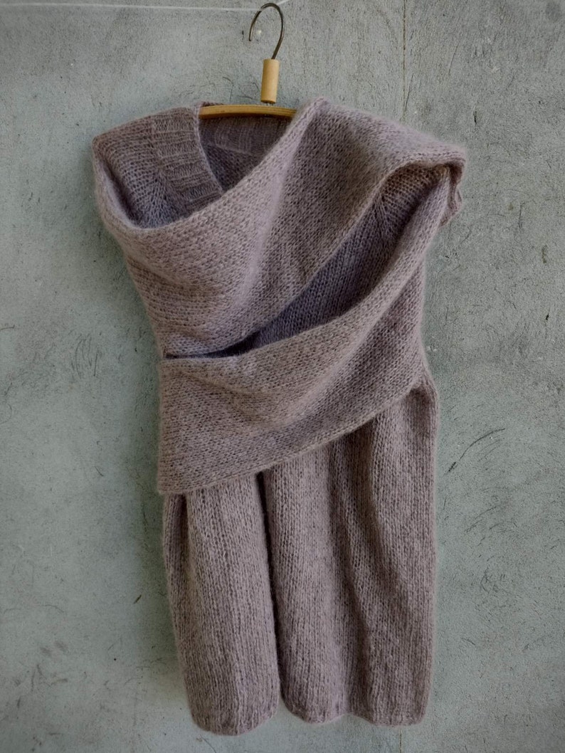 Pu&ograve; includere: Un maglione di maglia morbida color malva con collo a lupetto e un design drappeggiato. Il maglione &egrave; appeso a un appendiabiti di legno su uno sfondo grigio.