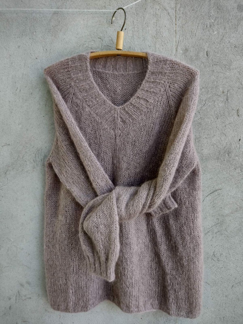 Pu&ograve; includere: Un maglione di lana morbido, di colore marrone chiaro, con scollo a V e maniche lunghe. Il maglione &egrave; appeso a un appendiabiti di legno.