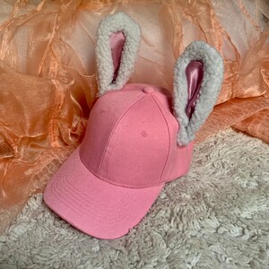 May include: A pink baseball cap with white fuzzy bunny ears. The cap has a curved brim and a white stitching detail around the brim.