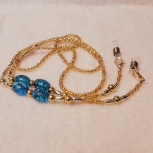 May include: A gold and silver beaded eyeglass chain with two turquoise blue glass beads. The chain has two clear plastic clasps for attaching to glasses.