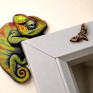 Pode incluir: Um acessório decorativo de parede com um camaleão vibrante em tons de verde e laranja com cauda enrolada, posicionado na borda de uma moldura de madeira cinza-claro, acompanhado por uma pequena mariposa castanha e bege no canto.