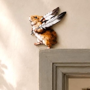 Könnte beinhalten: Ein braun-weißer Hamster-Wandaufkleber, der das Nagetier mit zwei großen Federn auf dem Rücken zeigt. Der Text "DOOR STORIES by Dear Prudence" ist in einem kreisförmigen Logo über dem Hamster auf der beigen Wandfläche aufgedruckt.