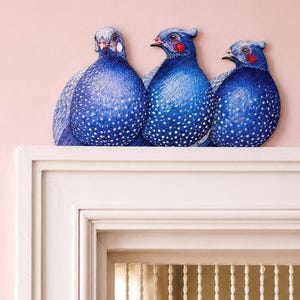 May include: Three decorative bird figures featuring vibrant blue plumage with white polka dot patterns. Each bird has a distinct head shape, small beak, and red accents near the eyes. The figures are arranged side by side on a white mantelpiece.