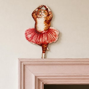 Könnte beinhalten: Ein Wandaufkleber zeigt eine orangefarbene getigerte Katze als Ballerina in einem roten Tutu mit erhobenen Armen. Rechts befindet sich ein kreisförmiges Logo mit dem Text DOOR STORIES by Dear Prudence und einem Schlüsselsymbol in der Mitte.