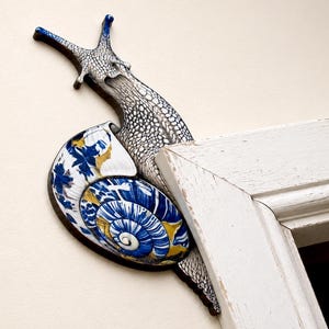 May include: A decorative snail wall ornament featuring a textured grey and white body with two antennae. The shell displays a vibrant blue, white, and yellow floral pattern. The snail is positioned to appear as if it is crawling around a white door frame.