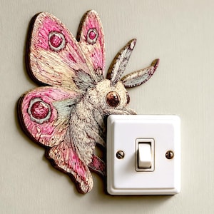 May include: A decorative wall decal featuring a moth with detailed pink and cream wings, positioned next to a standard white rectangular light switch. The moth has large dark eyes and textured wing patterns with prominent circular markings.
