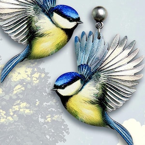May include: A pair of bluebird earrings with silver accents. The birds are painted in realistic detail with blue, yellow, and white colors. The earrings are made of wood and have a silver hook.